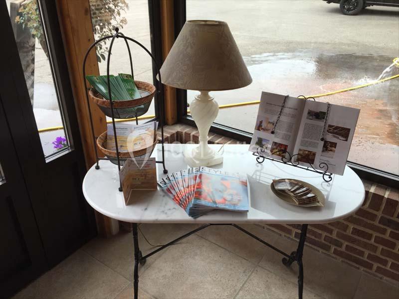 Table avec lampe, présentoir à brochures, magazines et dépliants, située près d'une fenêtre dans un espace lumineux.