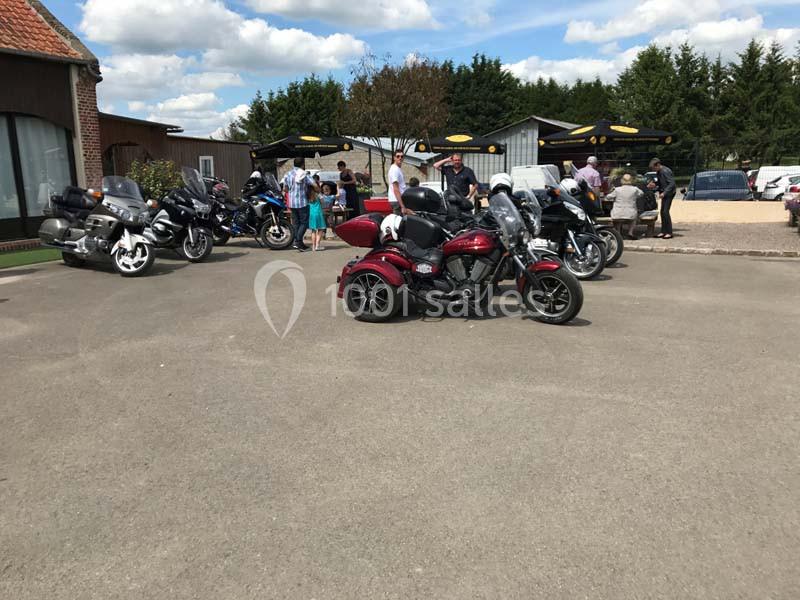 Plusieurs motos stationnées sur un parking devant un bâtiment, avec des personnes discutant à proximité.