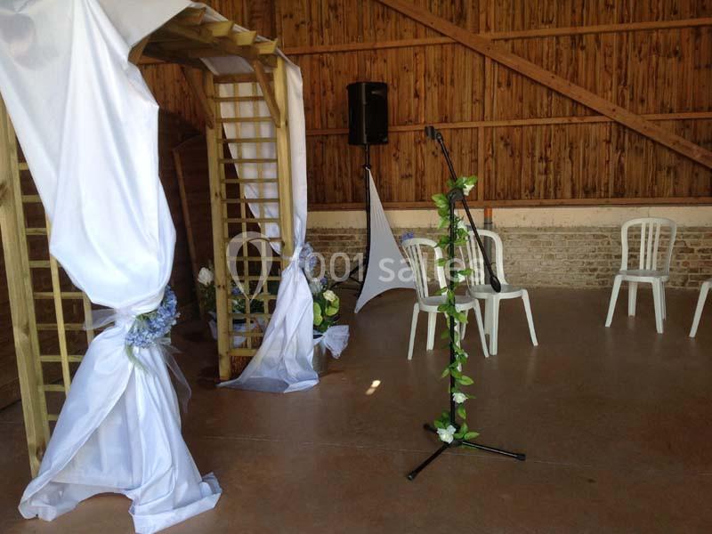 Arche en bois décorée de voilages blancs et de fleurs, avec un micro sur pied entouré de feuillage dans une salle en bois.
