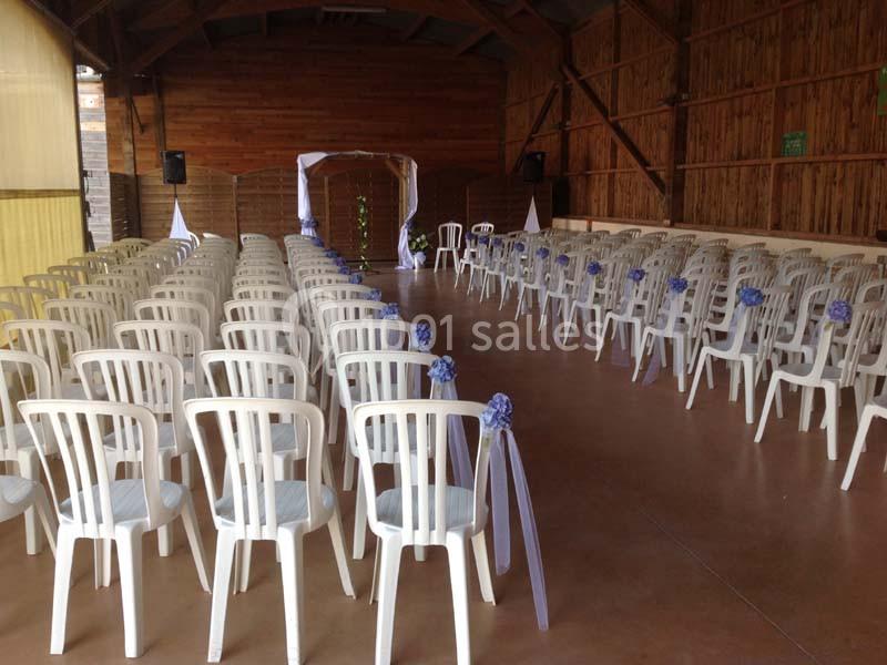Salle décorée pour une cérémonie, avec des rangées de chaises blanches ornées de rubans violets et une arche au fond.