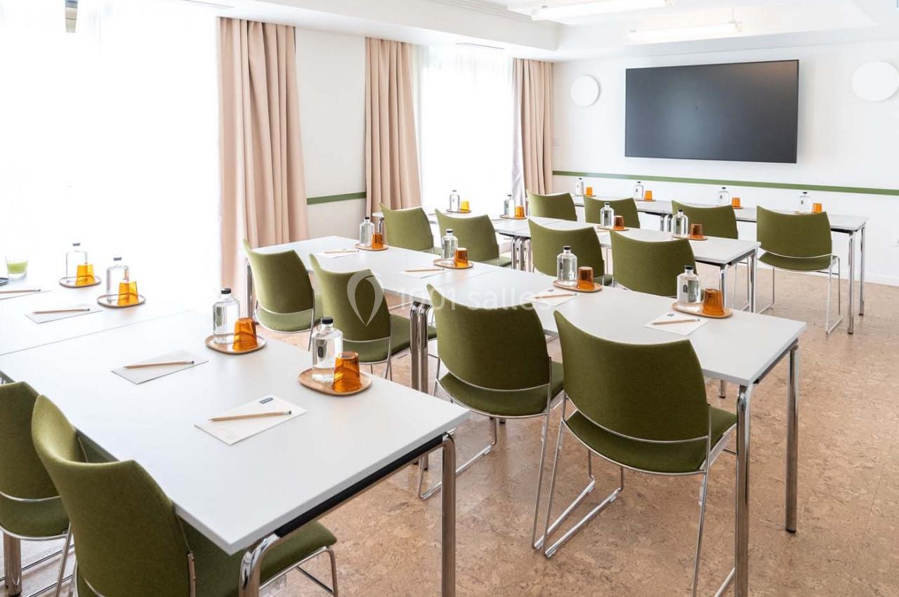 Salle de réunion lumineuse avec des tables blanches, des chaises vertes et un écran noir fixé au mur.