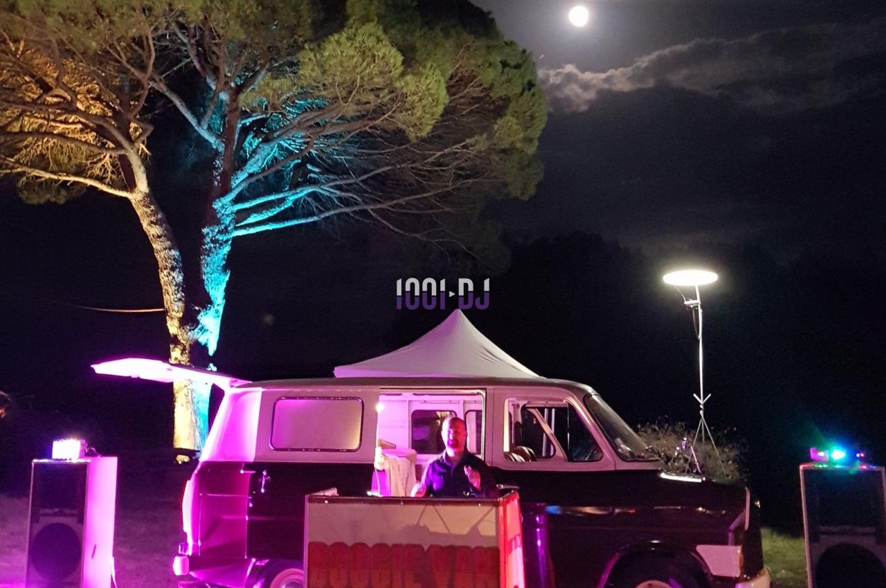 Un DJ mixe devant un van éclairé, entouré de haut-parleurs, sous un ciel nocturne avec une pleine lune.