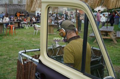 Un DJ mixe à l'extérieur depuis un stand aménagé dans un véhicule, avec des tables et des personnes en arrière-plan.