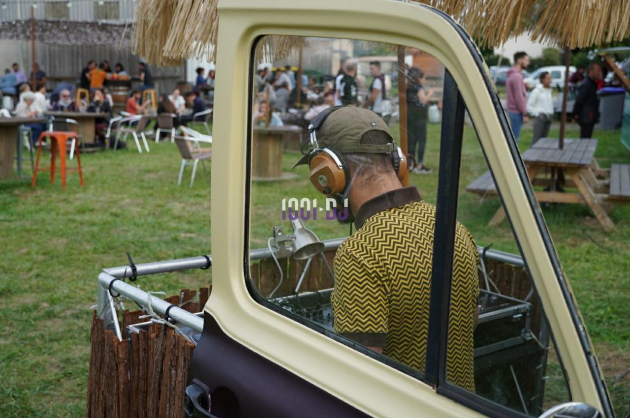Un DJ mixe à l'extérieur depuis un stand aménagé dans un véhicule, avec des tables et des personnes en arrière-plan.