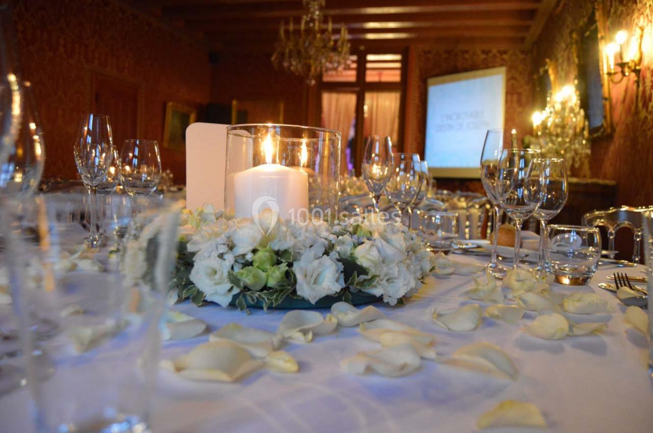 Table élégamment dressée avec une bougie entourée de fleurs blanches, pétales éparpillés, dans une salle ornée de lustres.