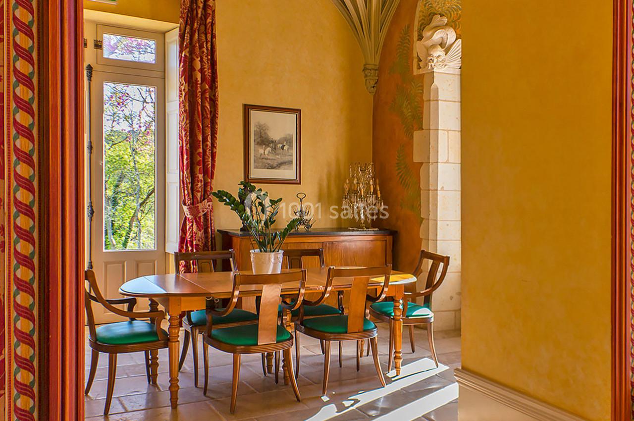 Salle à manger lumineuse avec table en bois, chaises à assise verte, murs jaunes et grandes fenêtres donnant sur un jardin.