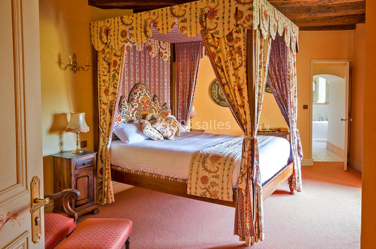 Lit à baldaquin avec tentures colorées dans une chambre chaleureuse aux murs orangés et poutres apparentes.