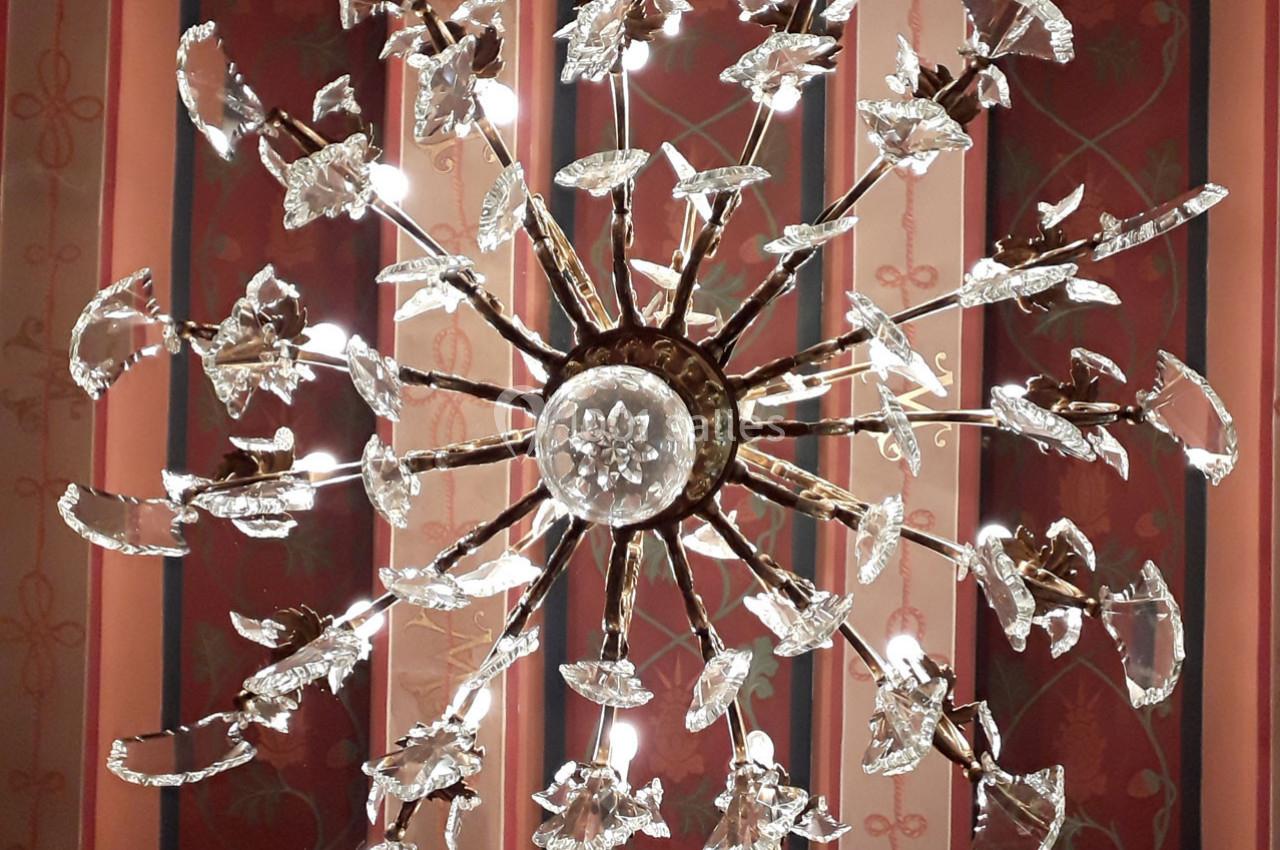 Lustre en métal doré orné de cristaux en forme de fleurs, suspendu devant un mur à motifs rouges et dorés.