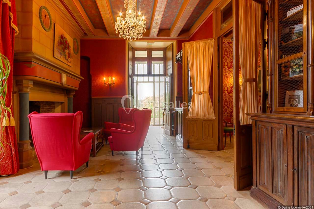 Salon élégant avec fauteuils rouges, cheminée, lustre et portes vitrées ouvertes sur un balcon avec vue extérieure.