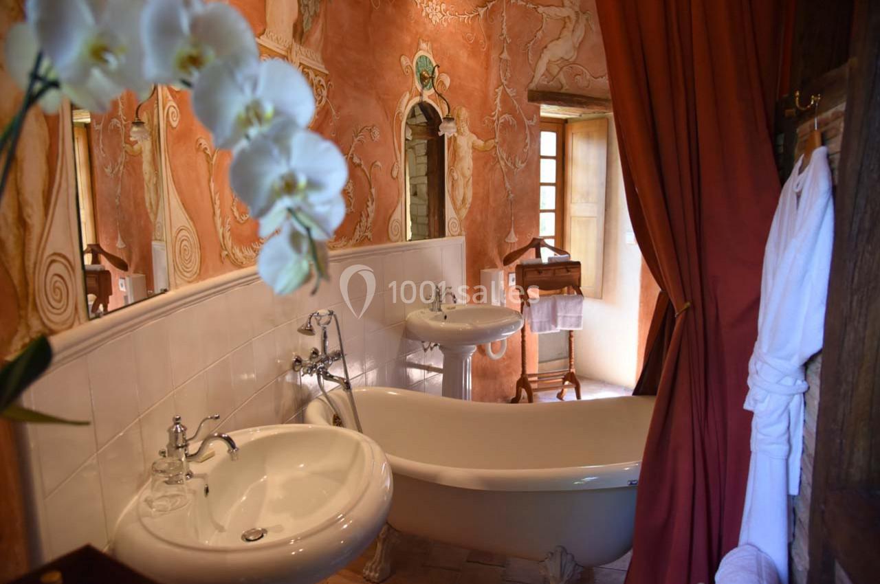 Salle de bain avec baignoire sur pieds, lavabo, mur peint en tons chauds et orchidée blanche au premier plan.