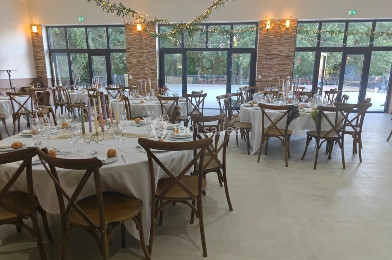 Salle de réception décorée avec des tables rondes dressées, des chaises en bois et des guirlandes lumineuses suspendues.