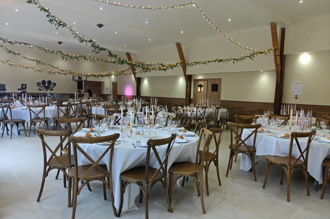 Salle de réception décorée avec des tables rondes dressées, chaises en bois et guirlandes lumineuses suspendues.
