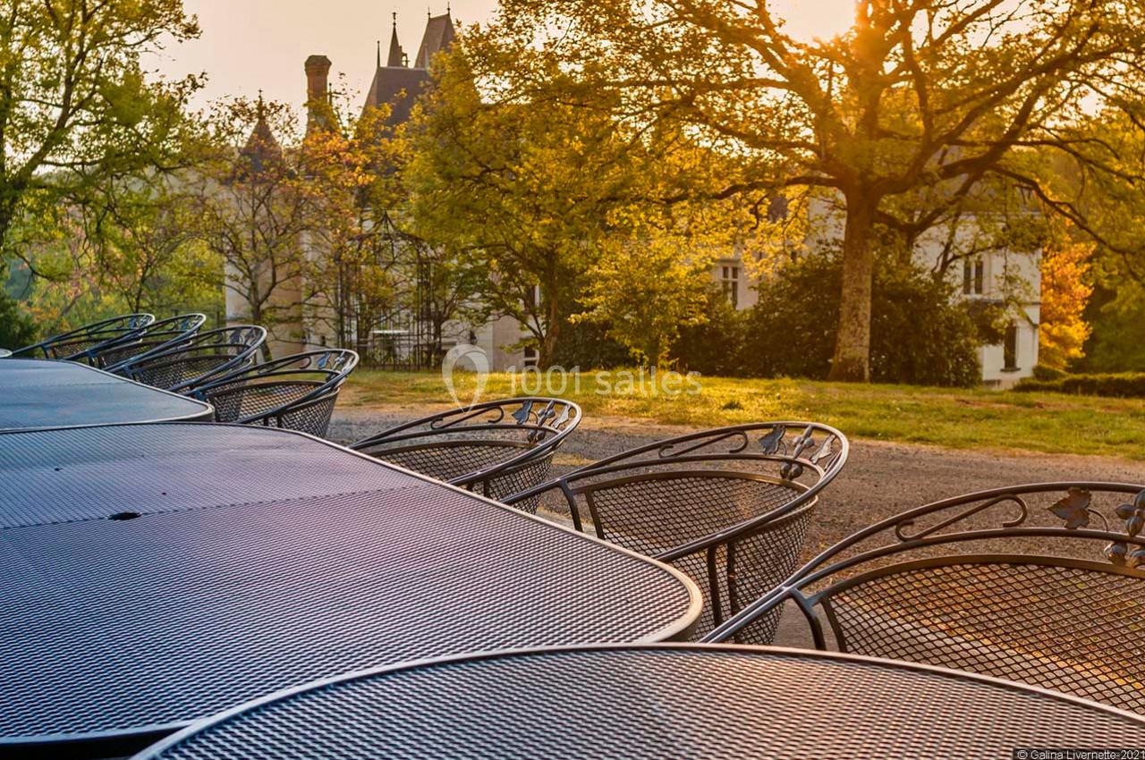Tables et chaises en métal alignées en extérieur, avec un château et des arbres éclairés par le soleil couchant en arrière…