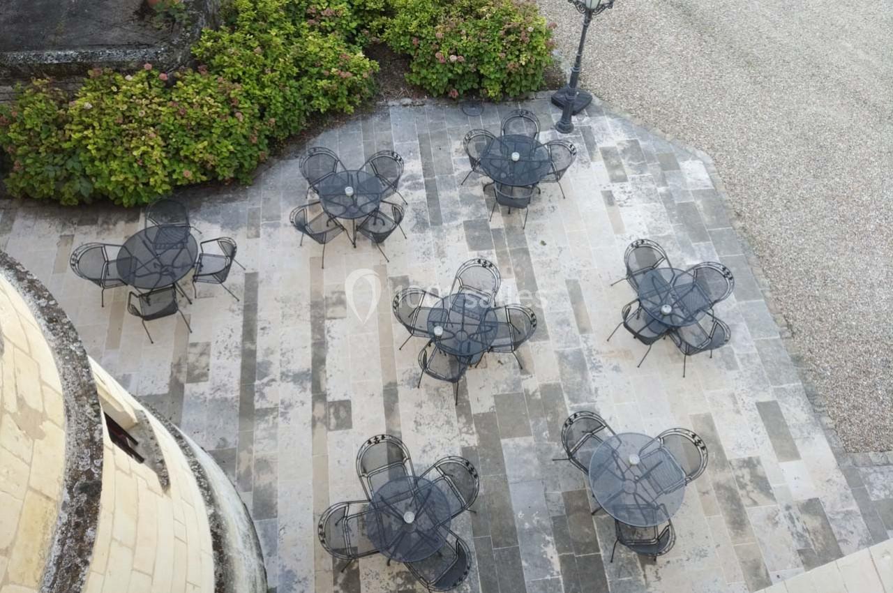Terrasse pavée avec plusieurs tables et chaises en métal noir, entourée de buissons et d'un lampadaire.