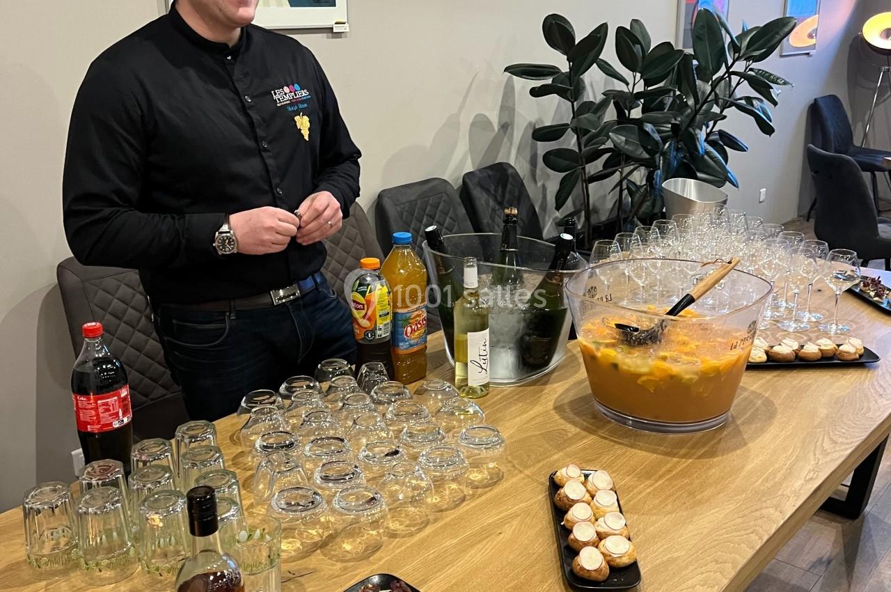 Un homme derrière une table garnie de boissons, verres, amuse-bouches et un grand saladier de cocktail.