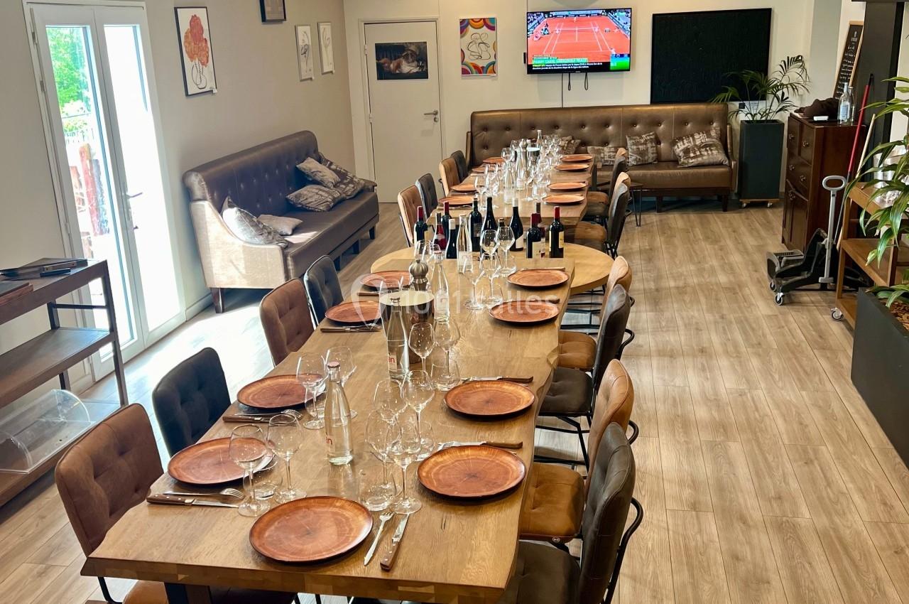 Table en bois dressée avec assiettes et verres, entourée de chaises, dans une salle lumineuse avec canapé et télévision…