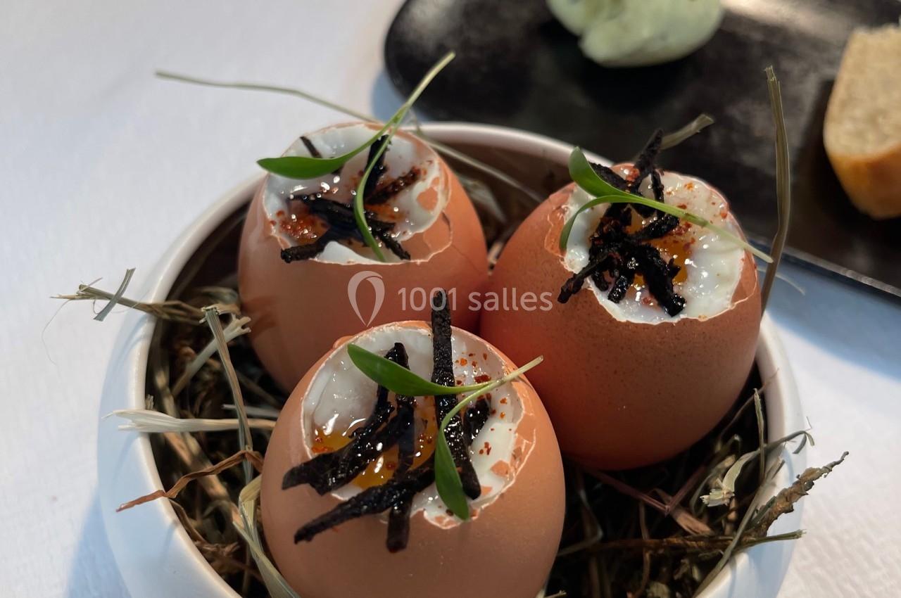 Œufs présentés dans des coquilles, garnis de crème et truffes, disposés sur un lit de foin dans un bol blanc.