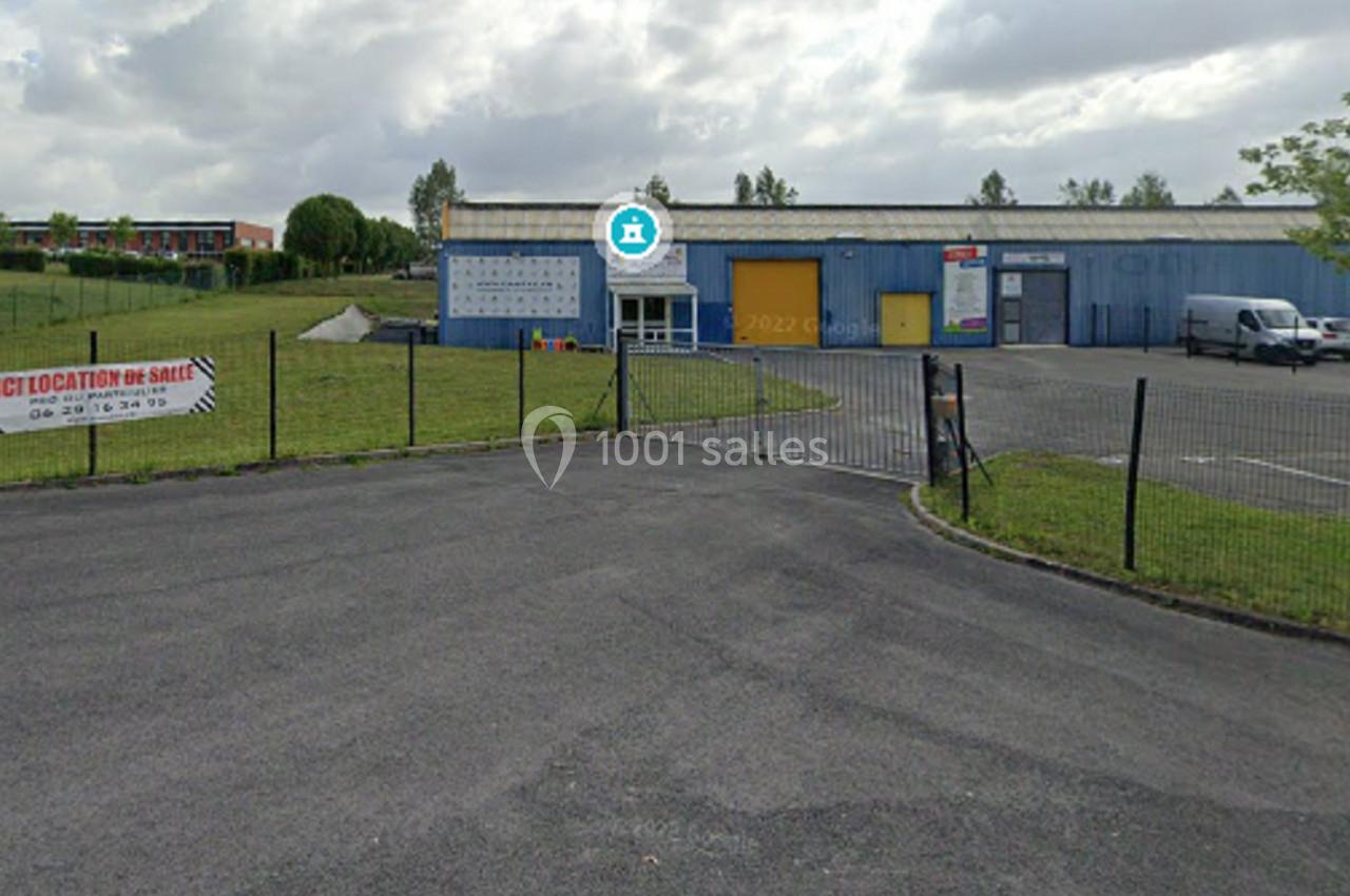 Entrée d'un parking clôturé devant des bâtiments industriels avec des panneaux publicitaires sur la clôture.