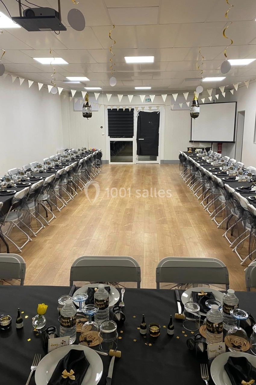 Salle décorée pour un événement, avec tables alignées, nappes noires, vaisselle élégante et guirlandes suspendues.