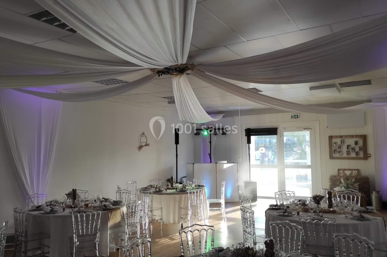 Salle de réception décorée avec des tables dressées, des chaises transparentes et des drapés suspendus au plafond.