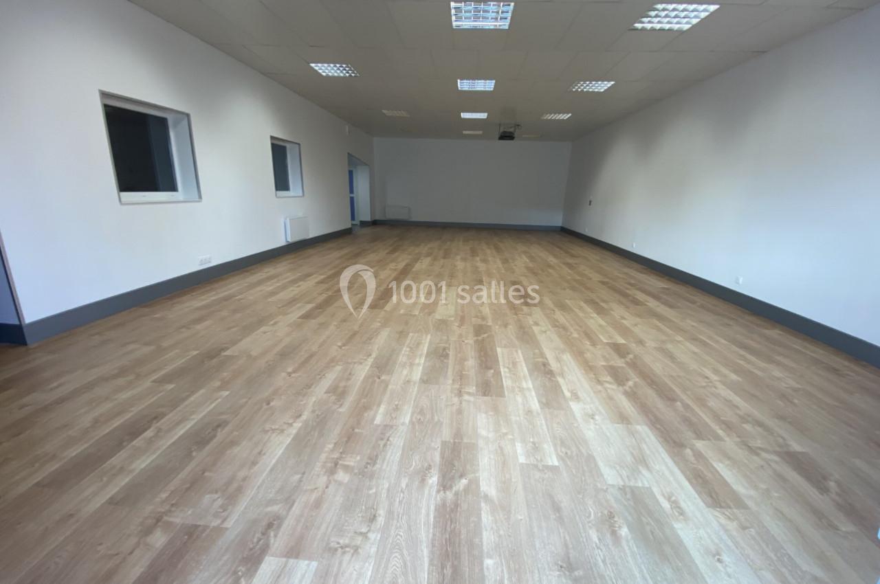 Salle vide avec parquet en bois clair, murs blancs, plafond avec éclairage encastré et fenêtres sur un côté.