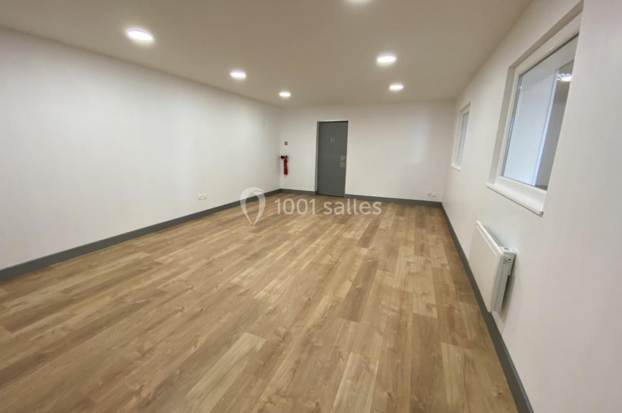 Salle vide avec parquet clair, murs blancs, éclairage encastré au plafond et une porte grise au fond.