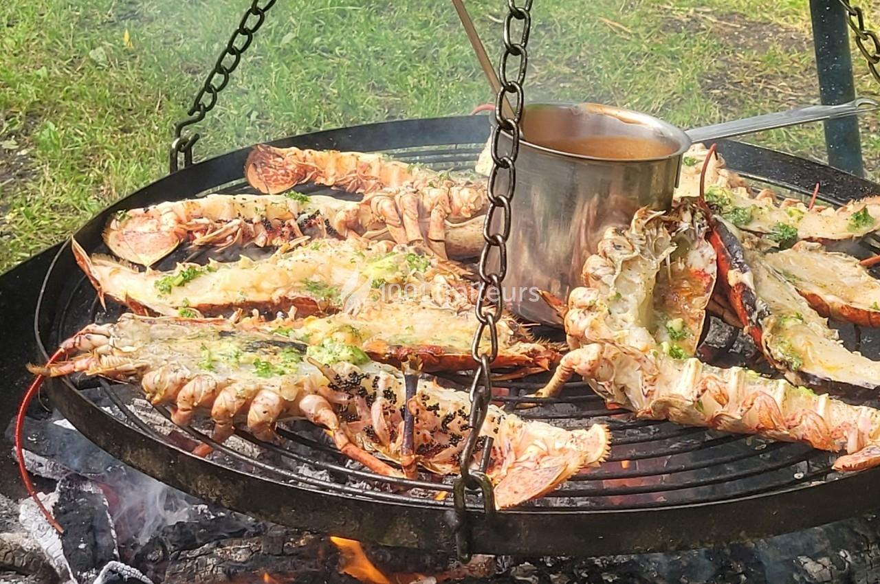 Homards grillés sur un barbecue suspendu au-dessus d'un feu de bois, avec une casserole en métal au centre.