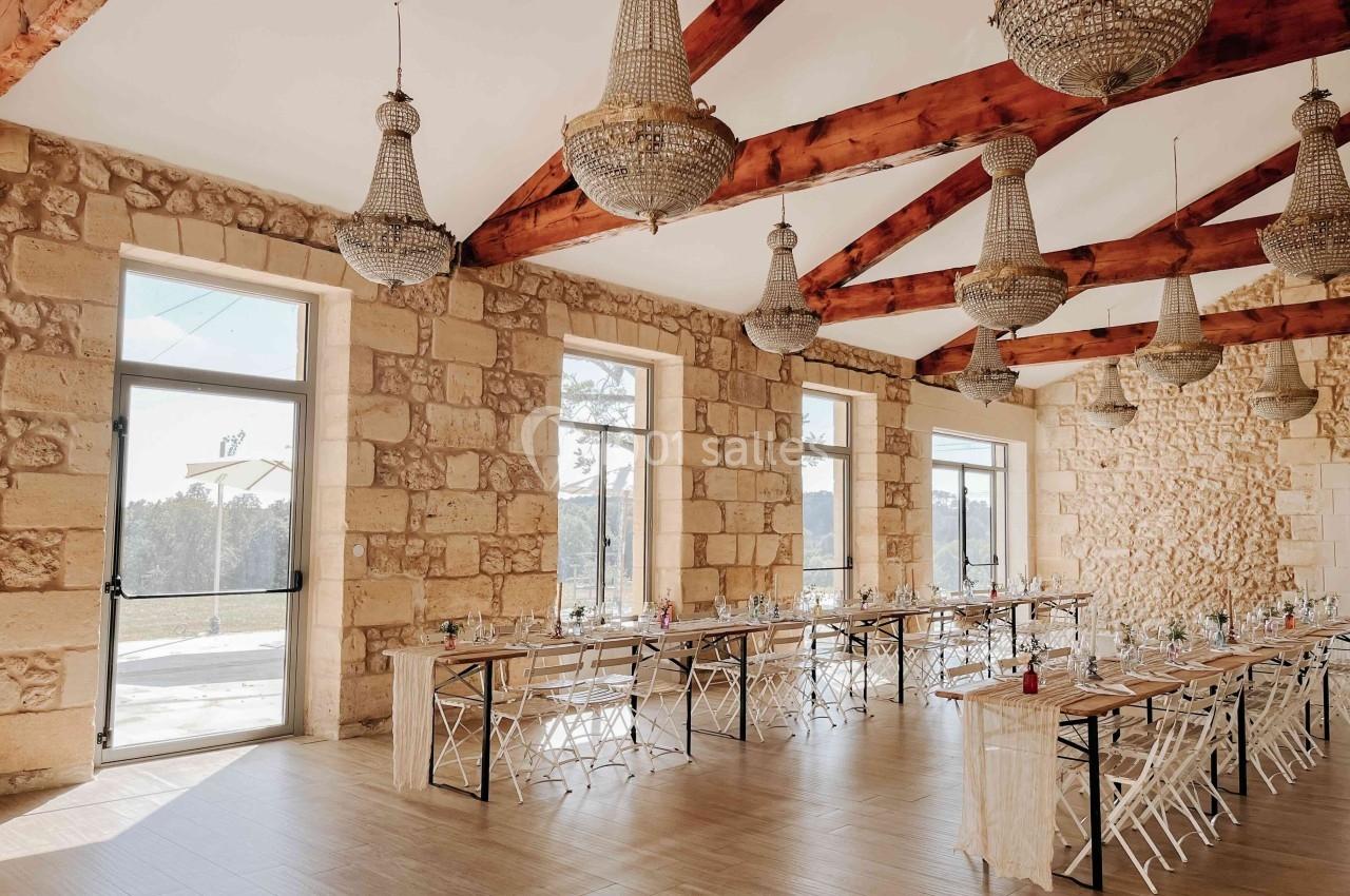 Salle lumineuse avec murs en pierre, poutres apparentes, lustres suspendus et tables alignées pour un événement.