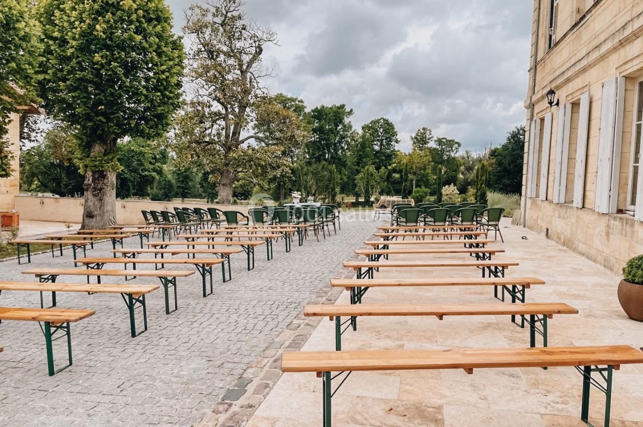Bancs en bois et chaises disposés en extérieur sur une terrasse pavée, entourée d'arbres et d'un bâtiment en pierre.