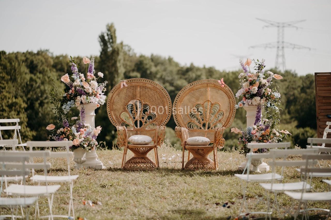 Deux fauteuils en rotin ornés de fleurs, disposés en extérieur sur une pelouse, entourés de chaises blanches.