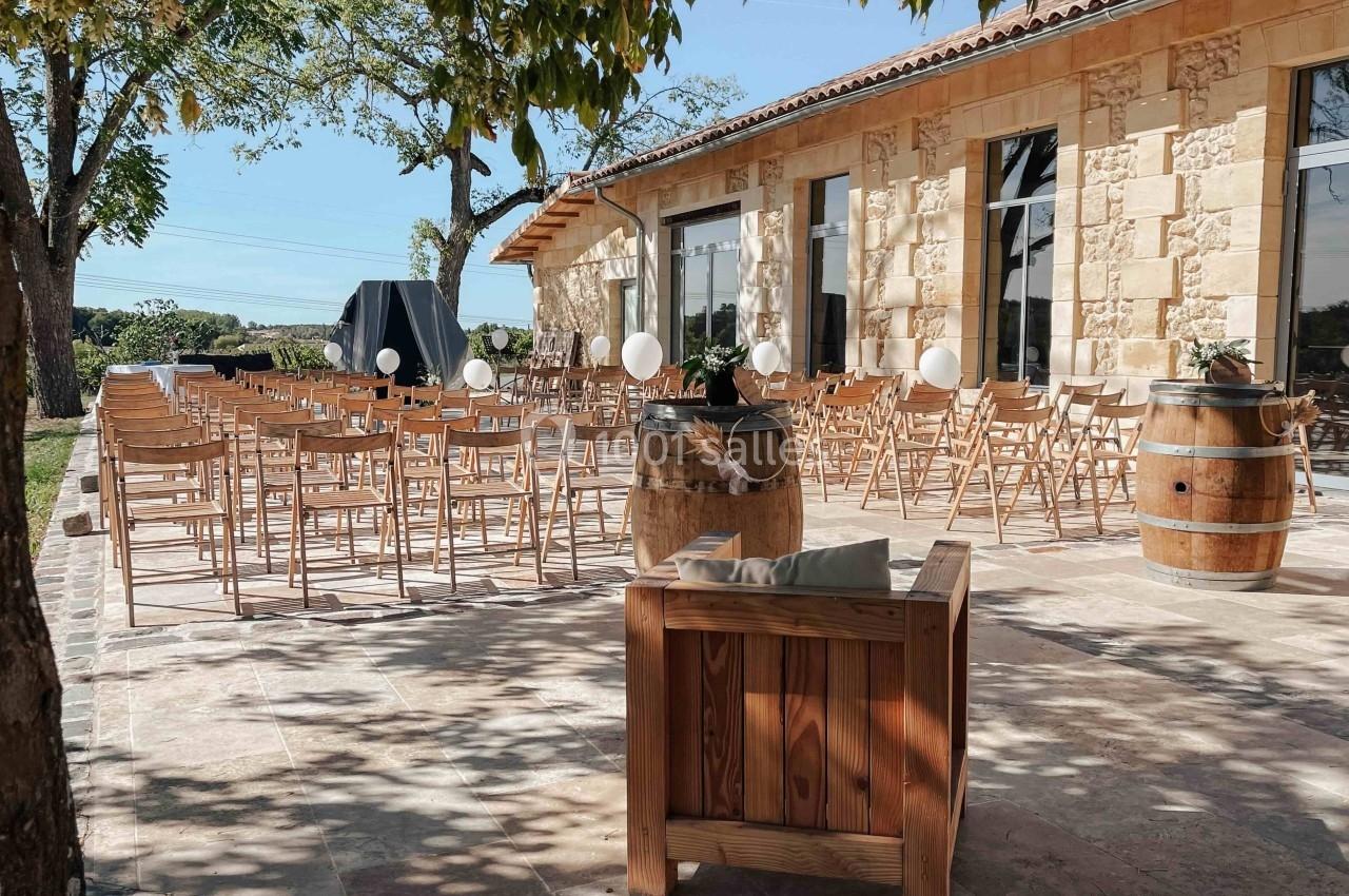 Chaises en bois disposées en rangs sur une terrasse en pierre, près d'un bâtiment en pierre, sous des arbres.