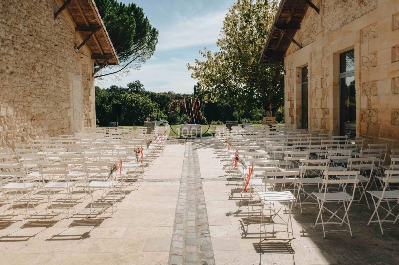 Chaises blanches disposées en rangées dans une cour en plein air, encadrée par deux bâtiments en pierre.