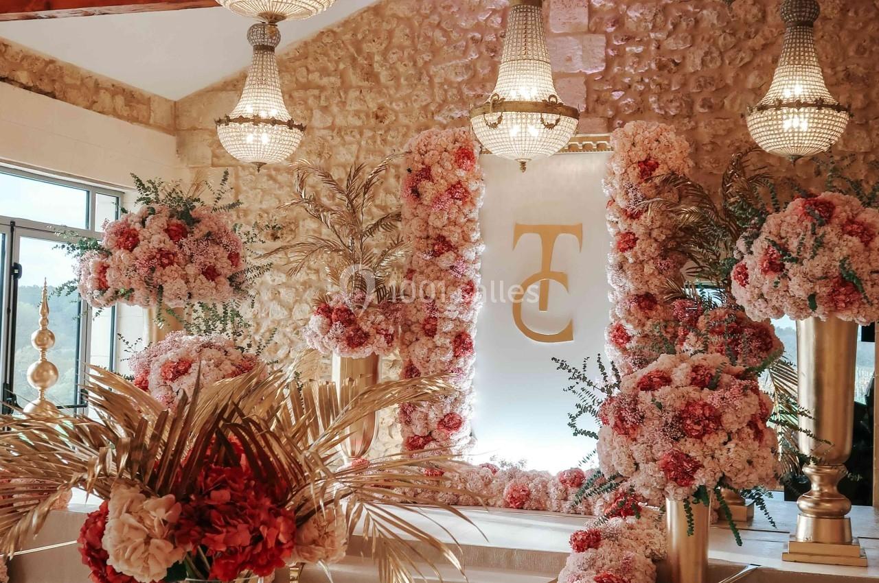 Salle décorée pour un événement avec lustres, fleurs roses et blanches, et un monogramme doré sur un panneau.