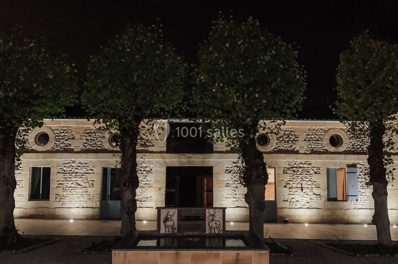 Façade d'un bâtiment en pierre éclairé la nuit, avec des arbres alignés et une petite fontaine au premier plan.