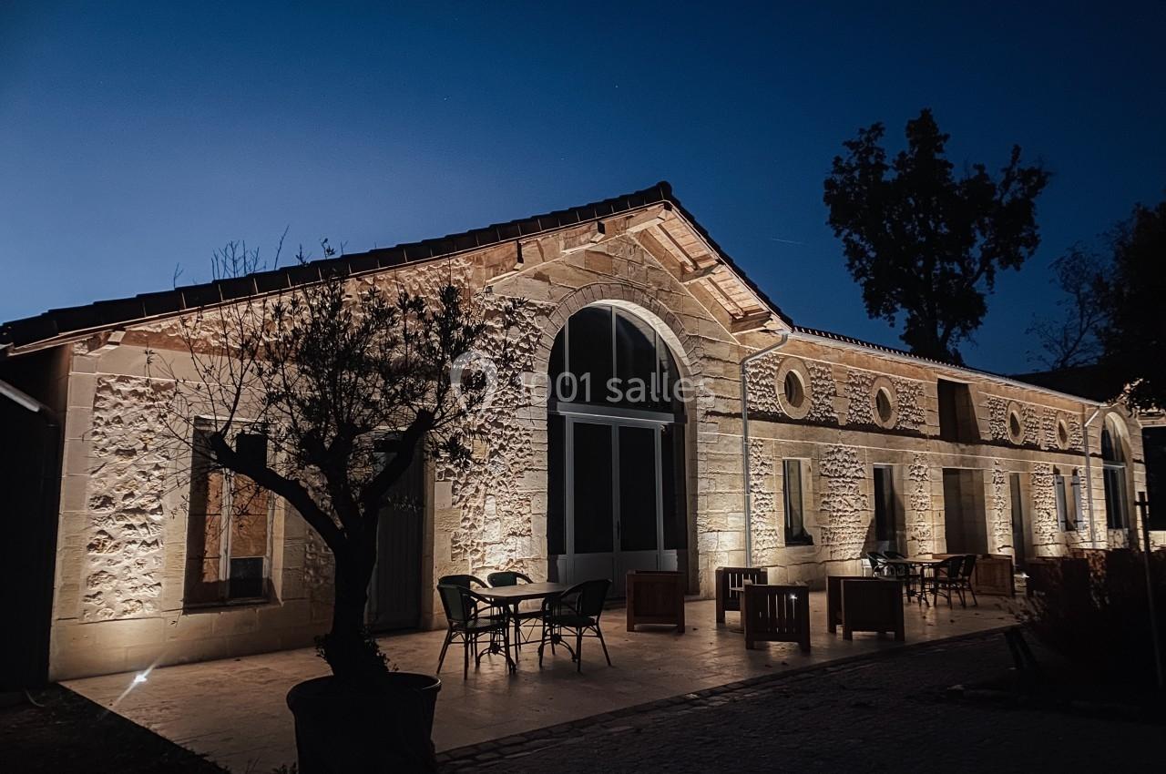 Façade en pierre d'un bâtiment éclairé la nuit, avec terrasse aménagée et arbre au premier plan.