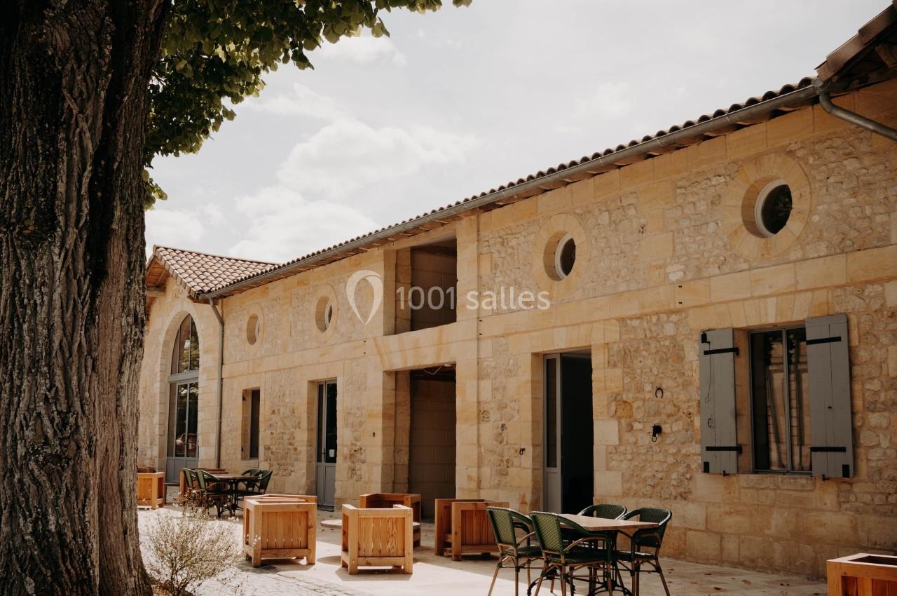 Façade en pierre d'un bâtiment avec terrasse aménagée de tables et chaises sous un arbre.
