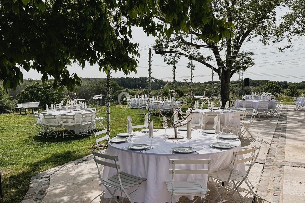 Tables rondes dressées avec nappes blanches et chaises pliantes dans un jardin verdoyant sous des arbres.