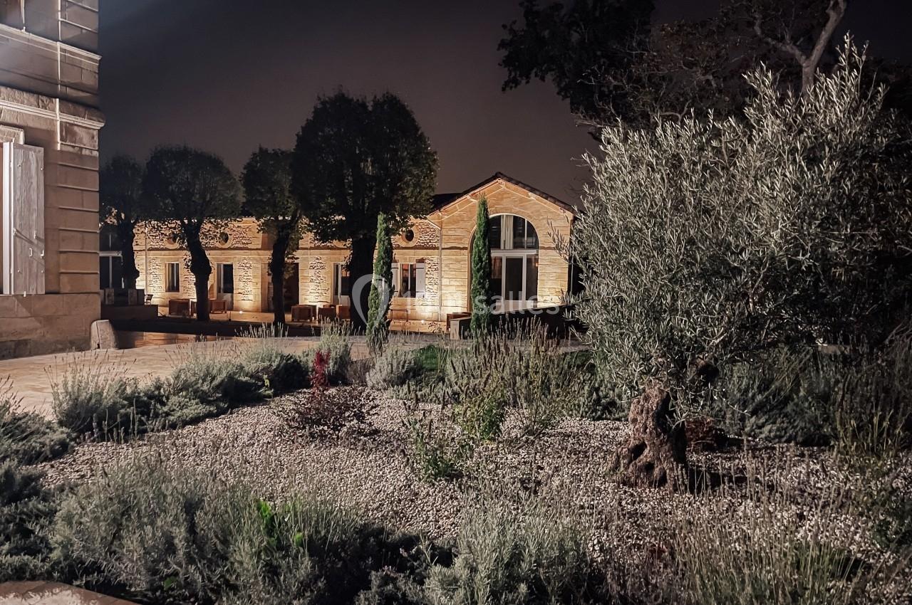 Jardin avec plantes méditerranéennes, éclairé la nuit, devant des bâtiments en pierre et des arbres taillés.
