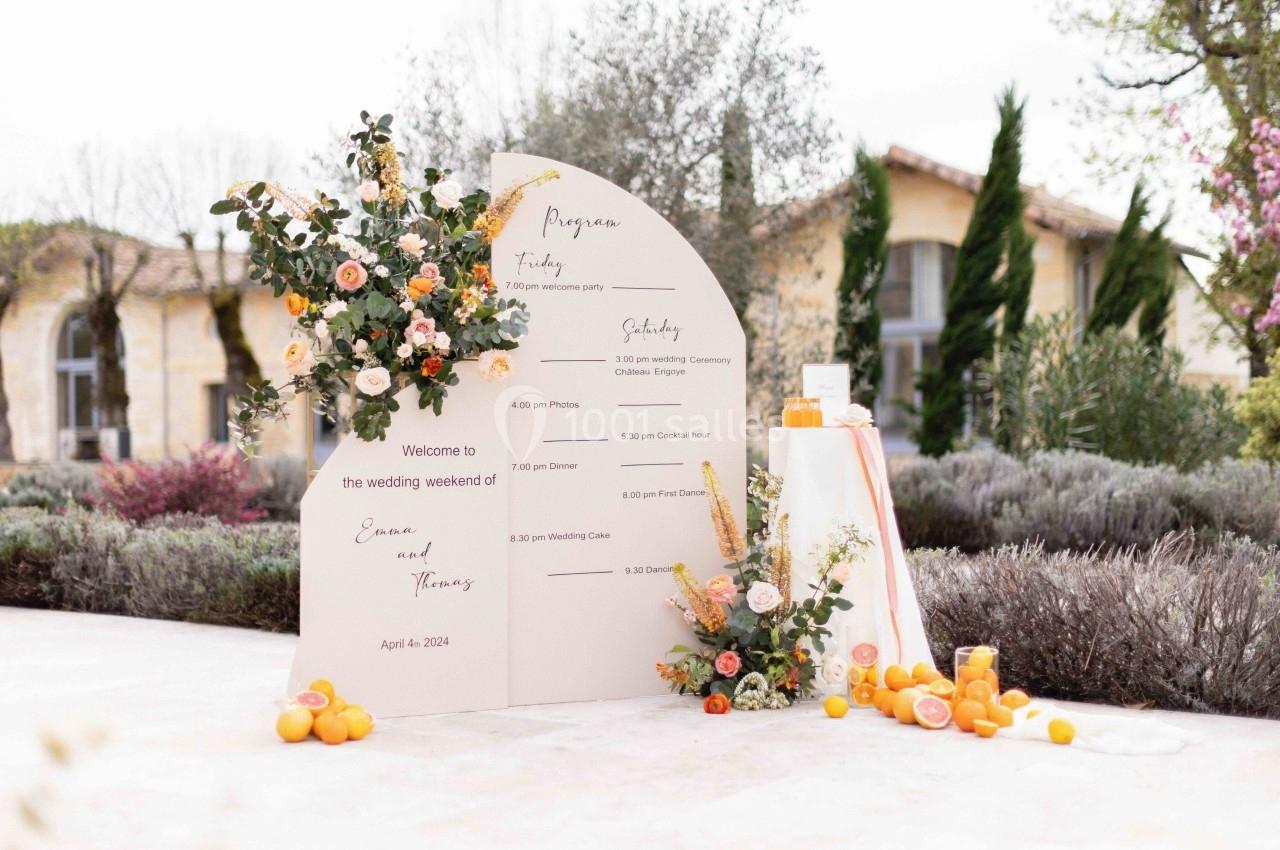 Panneau de programme de mariage décoré de fleurs et d'agrumes, placé dans un jardin avec des bâtiments en arrière-plan.