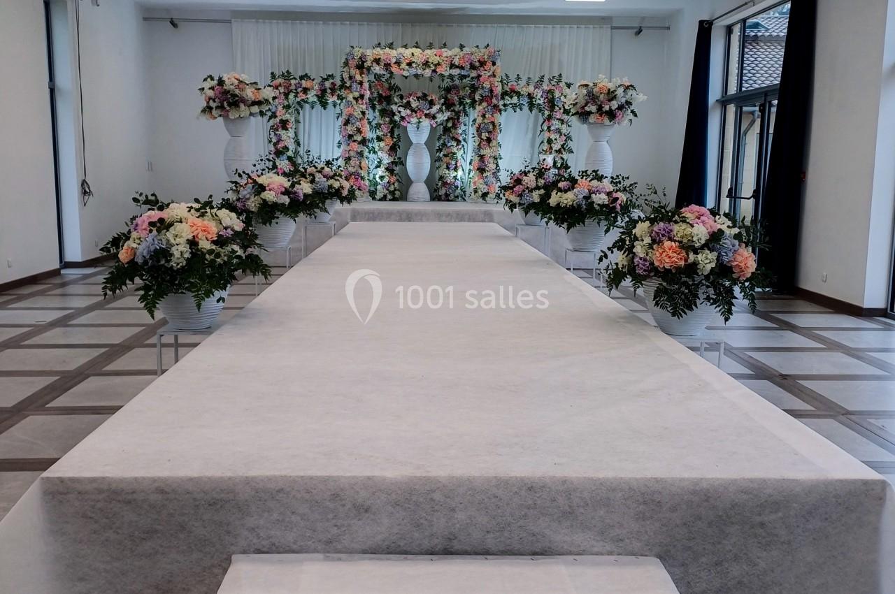 Allée centrale décorée de fleurs menant à une arche florale dans une salle de réception lumineuse.