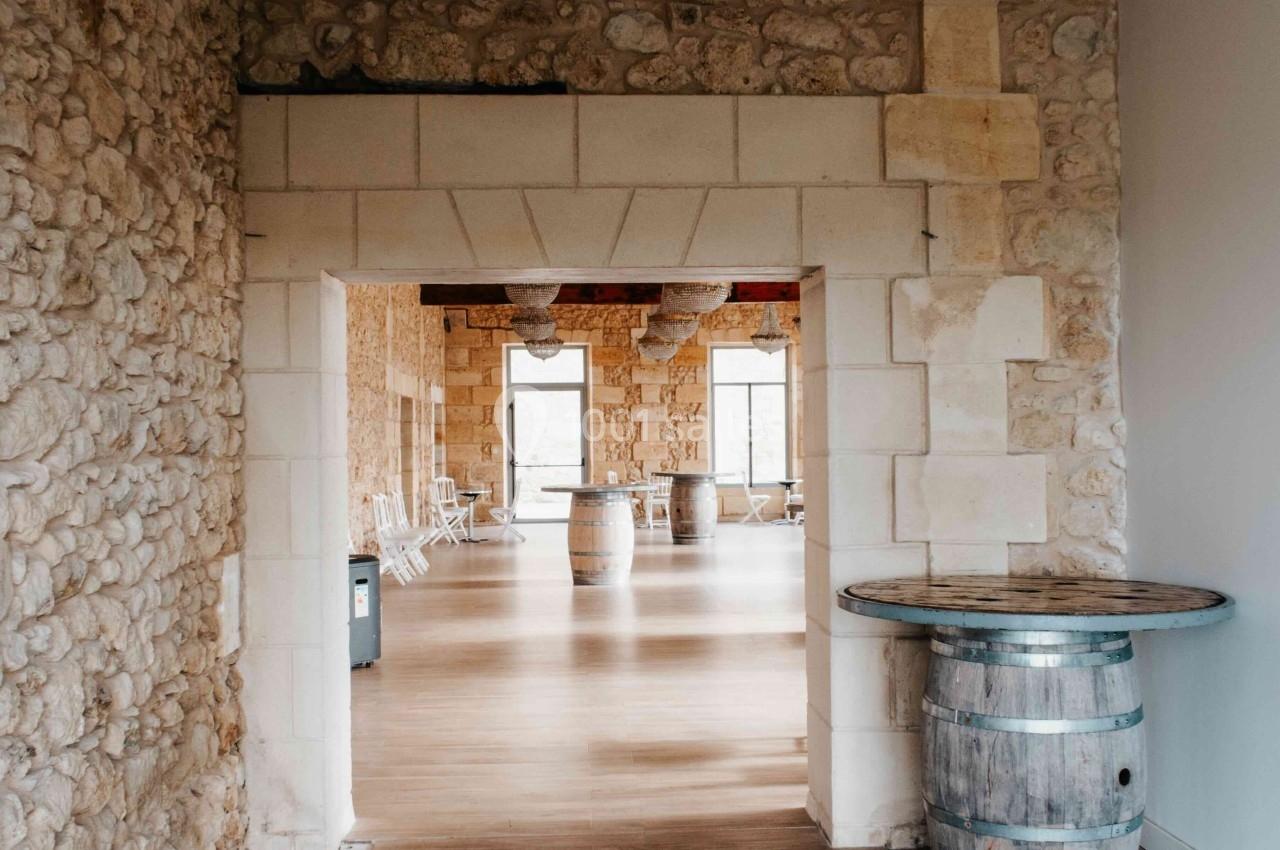 Entrée d'une salle lumineuse avec murs en pierre, parquet en bois et tonneaux servant de tables.