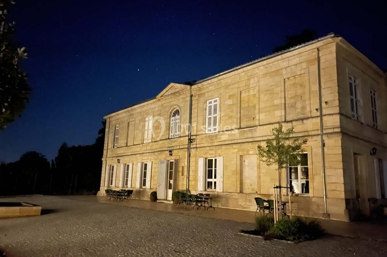 Façade d'un bâtiment en pierre éclairé la nuit, avec des tables et des chaises disposées sur une cour pavée.