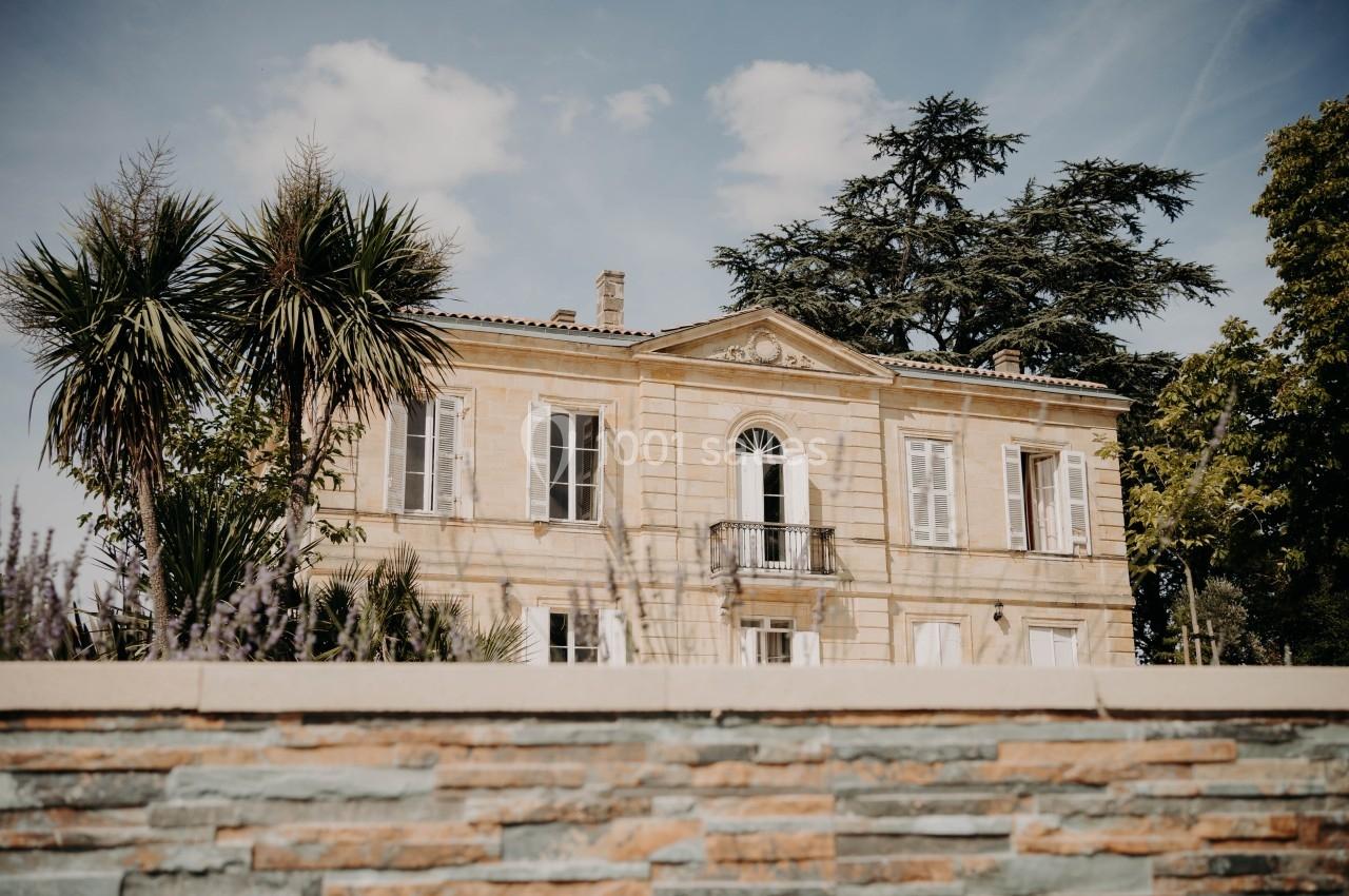 Façade d'une maison en pierre avec balcon, entourée de palmiers et d'arbres, vue depuis un mur en pierre.