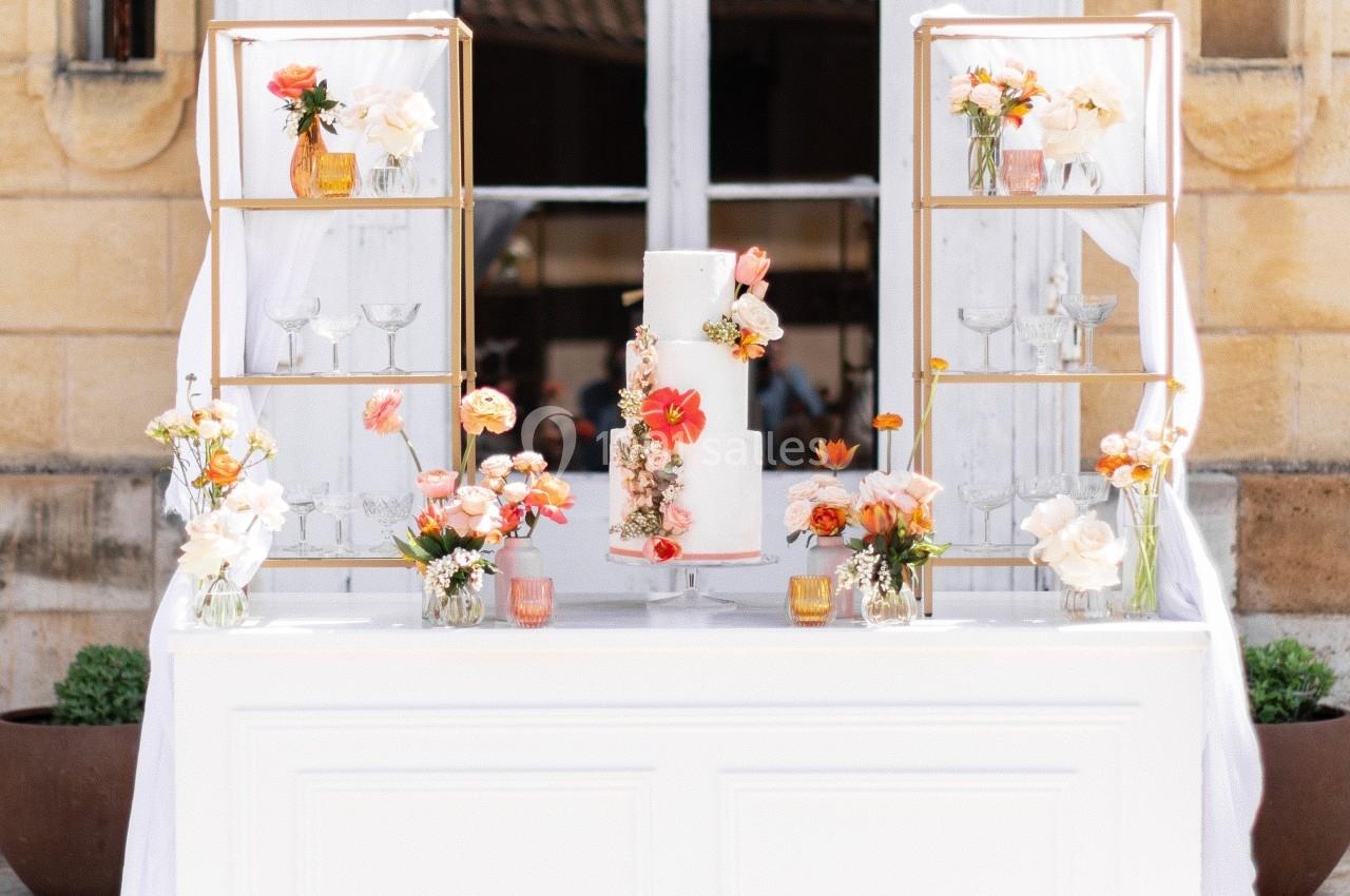 Table décorée pour un mariage avec un gâteau central, fleurs colorées et étagères dorées en arrière-plan.