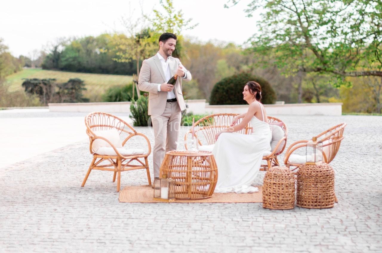 Un couple en tenue de mariage discute autour d'un mobilier en rotin sur une terrasse extérieure.