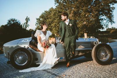 Une mariée en robe blanche et un marié en costume vert posent sur une voiture ancienne dans un cadre extérieur ensoleillé.
