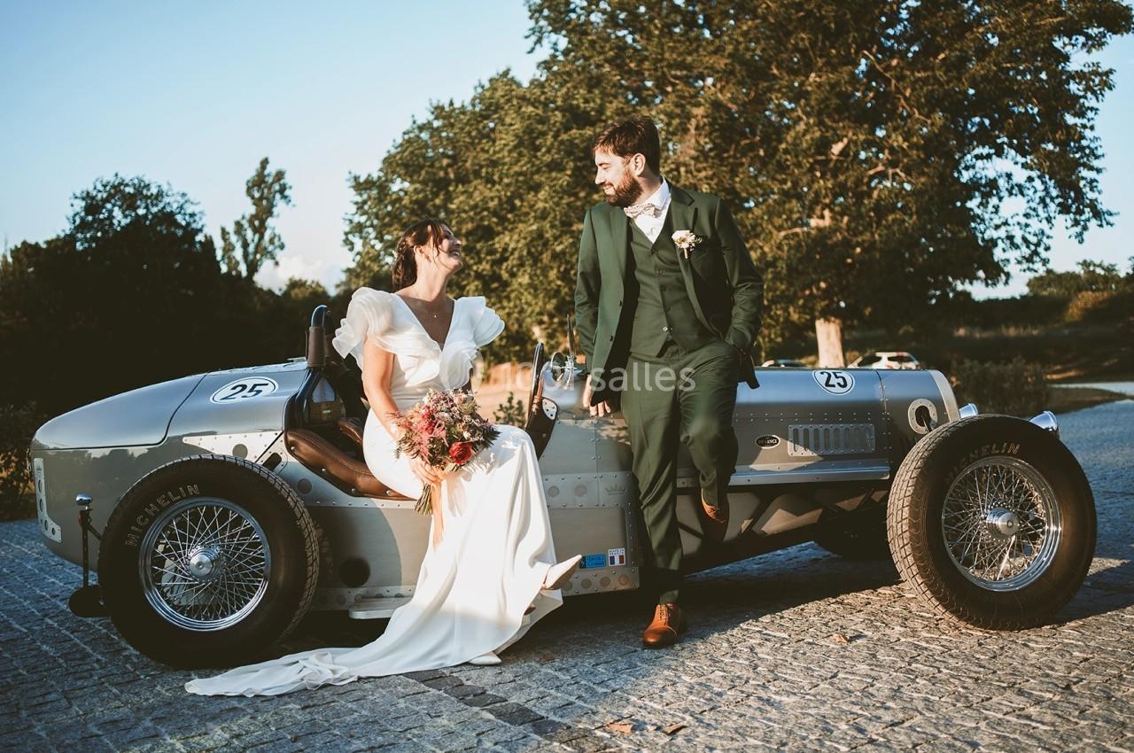 Une mariée en robe blanche et un marié en costume vert posent sur une voiture ancienne dans un cadre extérieur ensoleillé.