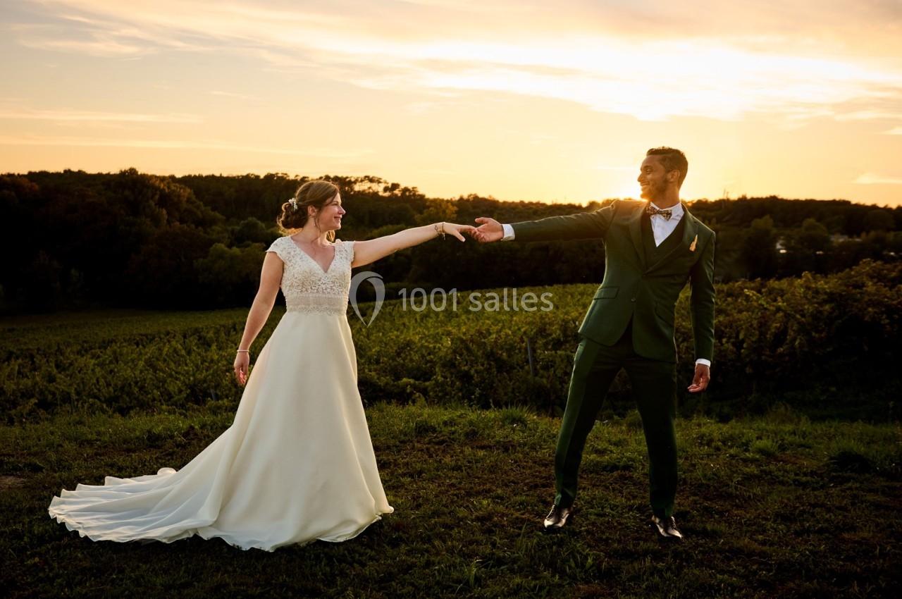 Un couple en tenue de mariage se tient la main dans un paysage champêtre au coucher du soleil.