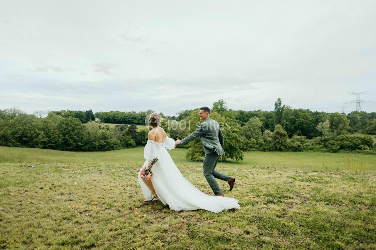 Un couple en tenue de mariage marche dans un champ verdoyant sous un ciel nuageux.