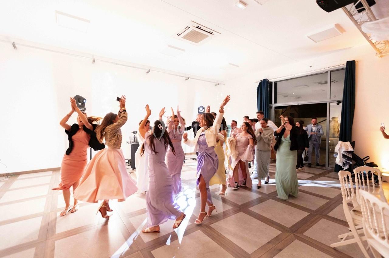 Des invités dansent joyeusement dans une salle lumineuse, vêtus de tenues élégantes et colorées.